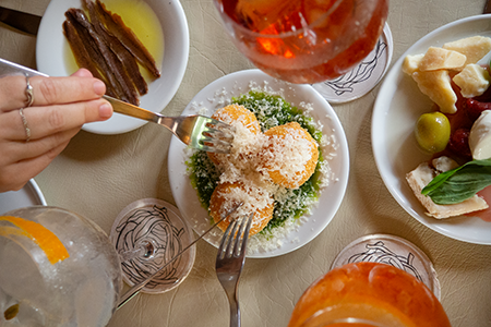 Mesa con platos de entradas y dos manos agarrando arancini con un tenedor